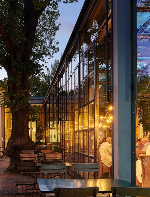 Glasfassade eines Restaurants bei Abenddämmerung, Terrassenmöbel und stimmungsvolle Beleuchtung schaffen eine einladende Atmosphäre.
