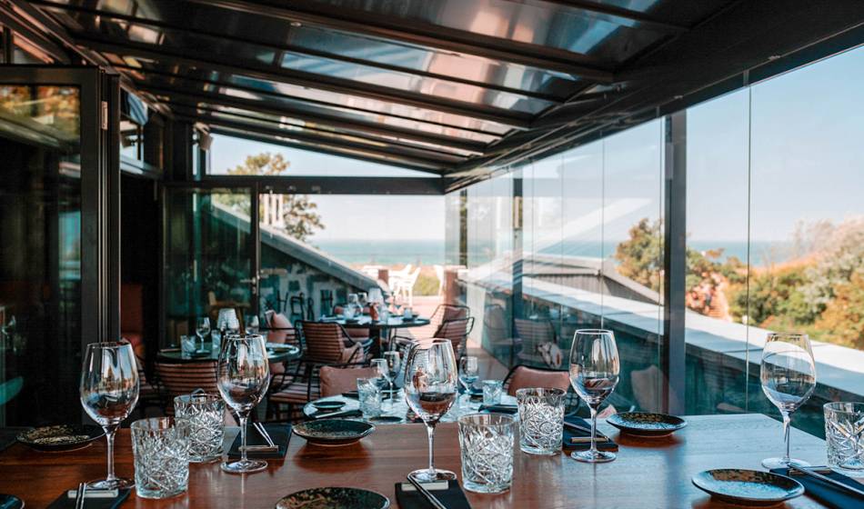 Wintergarten mit gedeckten Tischen, Gläsern und Besteck, umgeben von Glaswänden mit Blick auf das Meer im #herrhoshi.