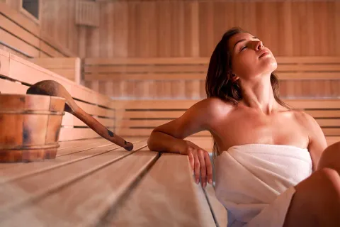 Eine Frau sitzt entspannt in der dampfenden Sauna, neben ihr stehen Schöpfkelle und Eimer.