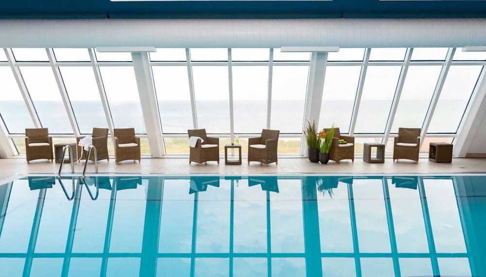 Heller Poolbereich im Wyn. Strandhotel Sylt mit Liegestühlen vor großen Fenstern, die einen Blick auf das Meer bieten.