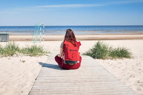 Frau mit rotem Rucksack sitzt am Ostseestrand auf Holzsteg mit Blick aufs Meer.