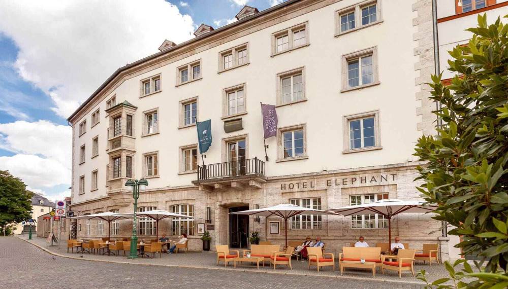 Außenansicht des Hotel Elephant Weimar mit Sitzmöglichkeiten, geöffneten Sonnenschirmen und Gästen.