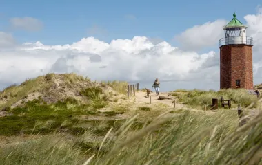 Ein Leuchtturm auf Sylt, umgeben von Dünen und einer Person mit Hund auf einem Spaziergang.