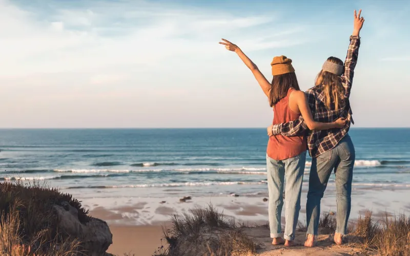 Zwei Freundinnen stehen Arm in Arm und blicken auf das Meer, friedliche Strandlandschaft im Hintergrund.