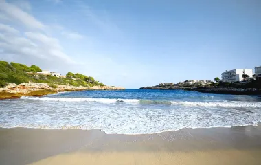 Strand mit feinem Sand, klarem Meer und umliegenden Häusern sowie grüner Natur unter einem strahlend blauen Himmel auf Mallorca.