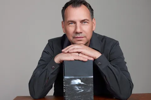 Autor Sebastian Fitzek sitzt an einem Tisch und stützt Hände und Kinn auf einem seiner Romane ab.