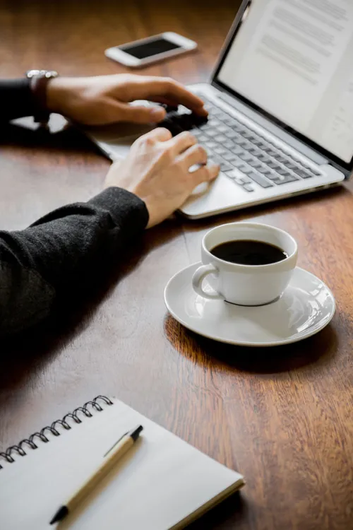Person tippt auf Laptop, daneben eine Tasse Kaffee, Notizblock mit Stift und Smartphone auf einem Holztisch in den Arcona Hotels & Resorts.