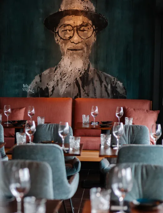 Restaurant mit gedeckten Tischen, Weingläsern und einer großen Wandmalerei eines Mannes mit Hut und Brille im Hintergrund.