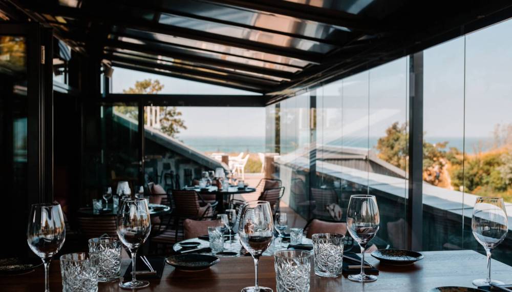 Restaurant #herrhoshi mit Glasfront und Meerblick, stilvolle Weingläser und Geschirr auf Holztischen in elegantem Ambiente.