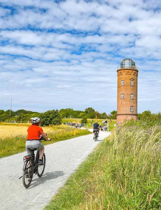 Fahrradfahrer auf einem Weg inmitten der Natur, der Leuchtturm steht im Hintergrund unter blauem Himmel.
