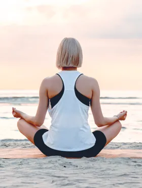 Eine Frau sitzt meditierend am Strand, der Blick auf das Meer gerichtet, während die Sonne aufgeht