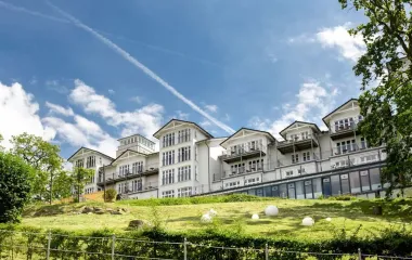 Außenansicht des Vju Hotels mit klassischer Architektur, großen Fenstern und Balkonen, gelegen auf einer grünen Anhöhe mit gepflegter Landschaft.