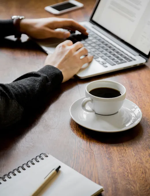 Person arbeitet am Laptop mit einer Tasse Kaffee und einem Notizbuch auf dem Tisch.