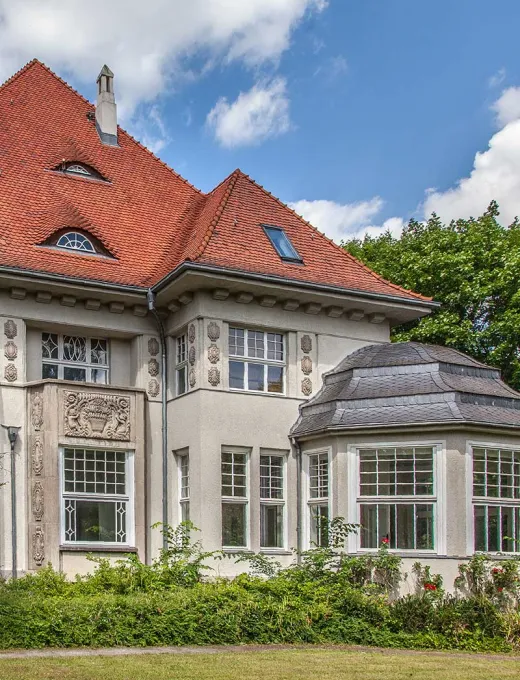 Historische Villa mit roten Ziegeldach, großen Fenstern und dekorativen Fassadenelementen.