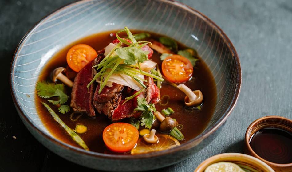 Eine Schüssel mit kräftiger Brühe, Rindfleisch, Pilzen, Tomaten und frischen Kräutern, kunstvoll arrangiert im #herrhoshi.