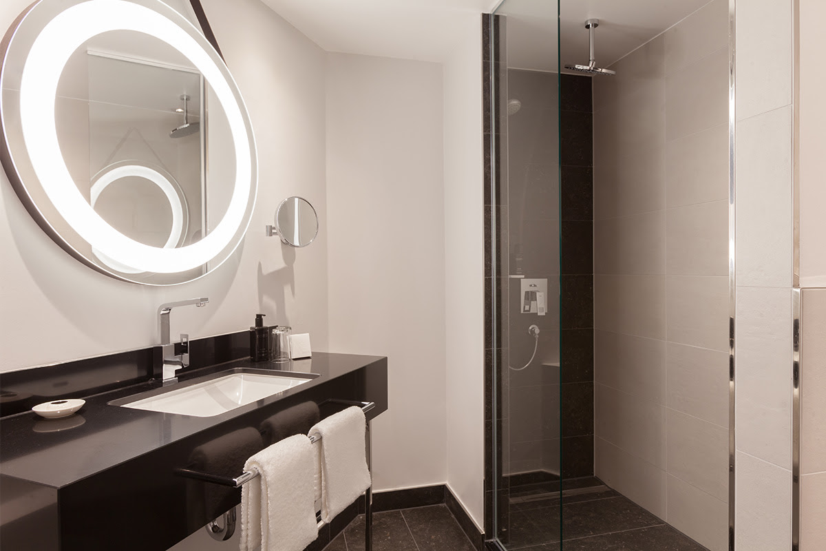 Modernes Badezimmer des Deluxe-Zimmers im Hotel Elephant Weimar mit Glasdusche, stilvollem Waschbecken und Spiegel.