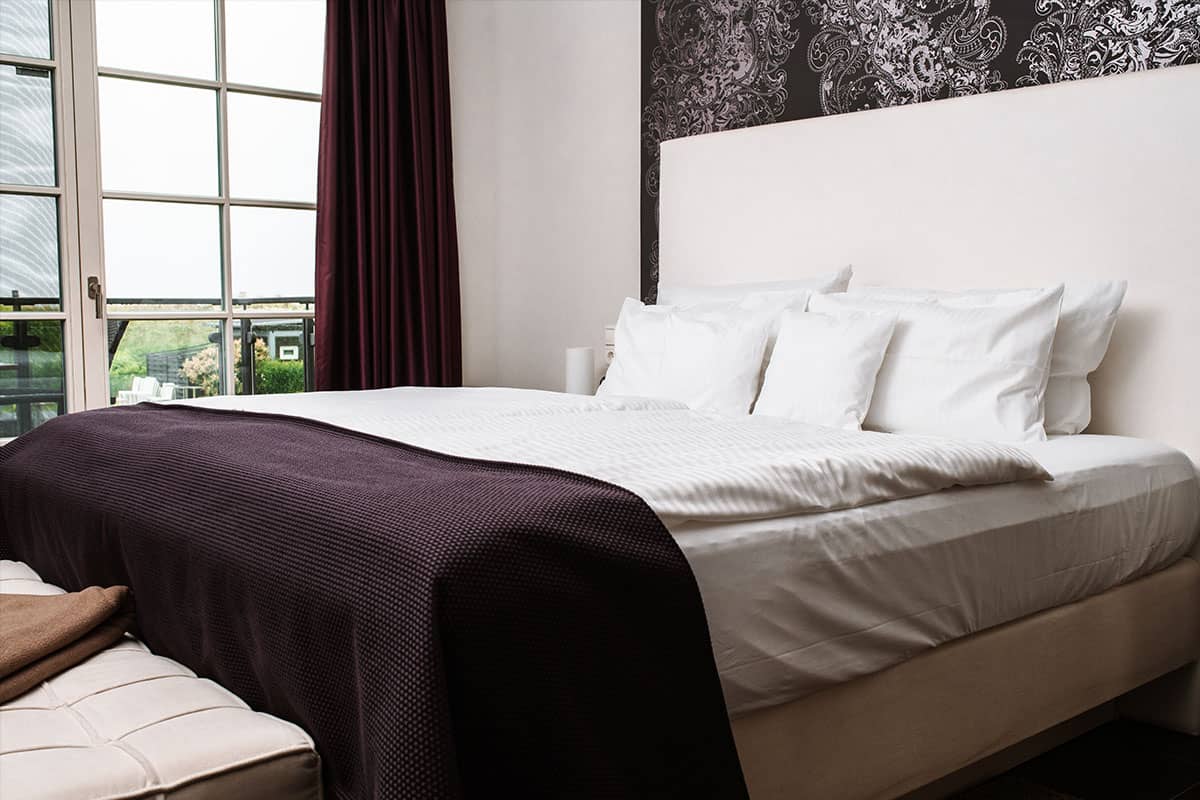 Großes Bett mit weißen Kissen und dunkelvioletter Tagesdecke in der Deluxe Suite im Hotel Künstlerquartier Seezeichen.