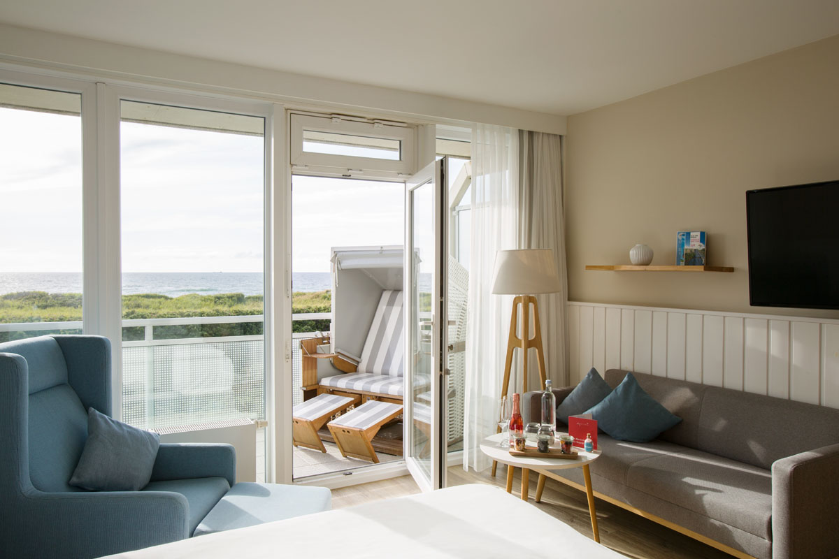 Wohnbereich im Zimmer Meerzeit des Wyn. Strandhotels mit Zugang zum Balkon, auf dem ein Strandkorb mit Aussicht auf das Meer steht.